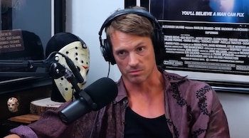 Un hombre rubio con auriculares y camisa morada en primer plano, con un micrófono. Detrás, una máscara de Jason Voorhees y un póster de Superman visible