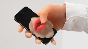 Una mano sostiene un teléfono móvil negro. Una estructura translúcida con labios rosados cubre parte de la pantalla. La mano lleva el puño de una camisa blanca