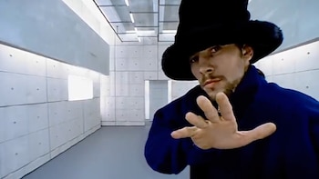 Jamiroquai, con sombrero oscuro y ropa azul y negra, caminando en una habitación gris con paredes de concreto y sofás negros