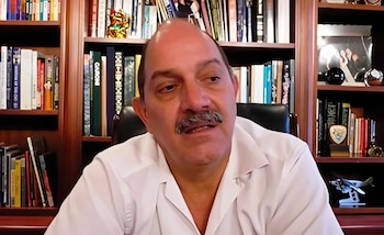 Hombre de tez clara con bigote, vistiendo camisa blanca, sentado frente a una estantería de madera con libros y adornos