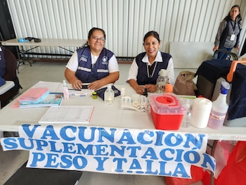 Dos trabajadoras con chalecos azules están tras una mesa con jeringas, recipientes médicos y una pancarta sobre vacunación, peso y talla