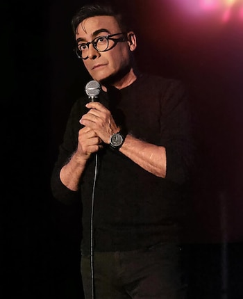 Adal Ramones, un hombre de mediana edad con gafas, de pie en un escenario oscuro, sosteniendo un micrófono con ambas manos