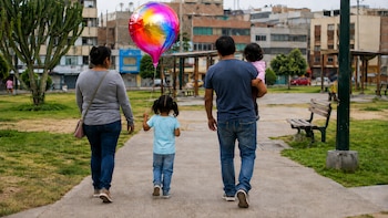 Familia de cuatro, dos adultos y dos niñas, camina de espaldas por un sendero de parque. Una niña pequeña sostiene un globo metálico multicolor. Hay césped y edificios