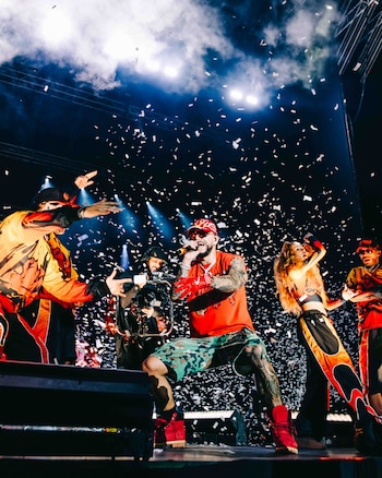 Ryan Castro, con camiseta roja, gorra y guantes rojos, canta en el escenario rodeado de confeti. A su alrededor hay bailarines y un camarógrafo, con luces y humo de fondo