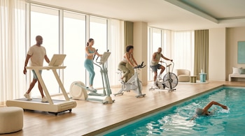 Gimnasio moderno con cinco personas ejercitándose: hombre en cinta, mujer en elíptica, mujer en bicicleta spinning, hombre en bicicleta de aire, y nadador en piscina cubierta.