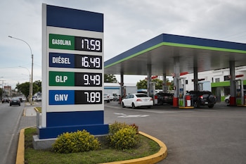 Un letrero electrónico muestra tarifas de combustibles junto a surtidores y vehículos estacionados en una gasolinera.