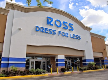 Fachada de una tienda Ross Dress for Less con el logo azul. La estructura es blanca con franjas azules y detalles en beige. Hay arbustos en la base y un cielo nublado