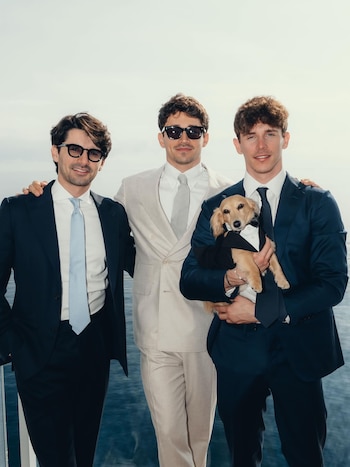 Tres hombres con traje y corbata posan frente al mar, uno de ellos sostiene un perro pequeño con esmoquin