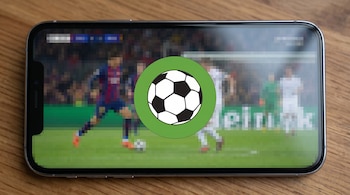 Primer plano de un teléfono móvil plateado en horizontal sobre una mesa de madera, mostrando un partido de fútbol pixelado y un logo de balón nítido.