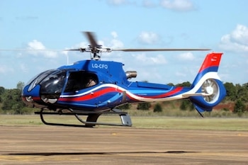 Helicóptero azul con rayas rojas y blancas, parcialmente en el aire sobre una pista de asfalto, con el rotor principal girando y árboles de fondo