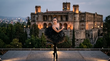 Una bailarina con tutú negro posa en puntas sobre una tarima frente a un castillo de piedra iluminado y árboles al anochecer.