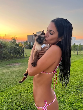 Silvina Escudero en bikini rosado y blanco besando a su gata siamés, Afrika, en un jardín verde con el cielo anaranjado del atardecer