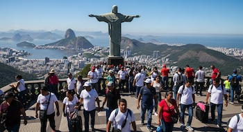 Estatua del Cristo Redentor, una multitud de turistas y familias, vista panorámica de Río de Janeiro con montañas y bahía, cielo azul claro.