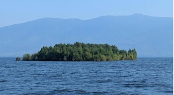 Isla cubierta de árboles en el centro de un cuerpo de agua con montañas azules y cielo despejado al fondo