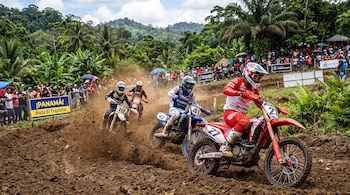 Pilotos de motocross con cascos y trajes de protección avanzan en motocicletas por un circuito de tierra con espectadores al fondo.