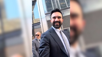 Un hombre con barba y traje oscuro sonríe ampliamente mientras camina al aire libre. Detrás, otro hombre con gafas de sol y traje