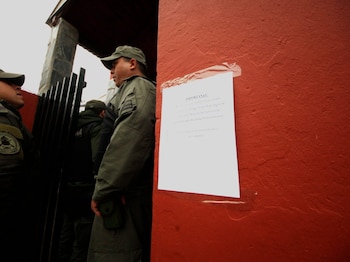 Tres personas con uniformes y gorras verde oscuro en una entrada. Una pared roja con un cartel blanco pegado que muestra texto impreso