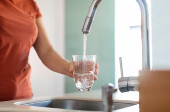 Una mano sostiene un vaso transparente debajo de un grifo de metal mientras se llena con agua en un fregadero