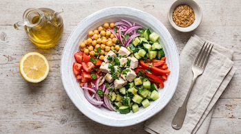 Plato con trozos de pollo, garbanzos, pepino, tomate, pimiento y cebolla, junto a medio limón, aceite, mostaza y un tenedor sobre madera.