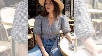 Una mujer con cabello oscuro y sombrero de paja luce una blusa cruzada de estampado vichy blanco y negro, jeans azules, y está sentada en una mesa de café con una taza.