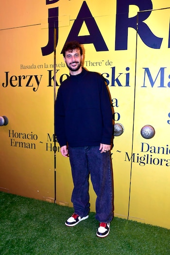 Hombre de barba sonríe, vistiendo sudadera azul oscuro y jeans, sobre césped artificial. Fondo amarillo con las letras "JARDÍN" y "Jerzy Kosinski"