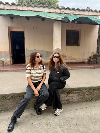 Dos mujeres con gafas de sol permanecen sentadas en el escalón exterior de una casa de fachada antigua y tejado de tejas