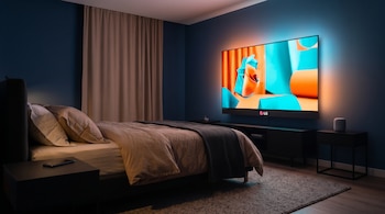 Un dormitorio a oscuras con una cama hecha, mesillas de noche y un televisor LG encendido con un anuncio colorido en la pared.
