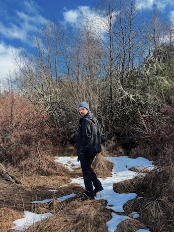 Hombre con gorro azul y gafas de sol, chaqueta oscura y pantalón, caminando por un sendero con nieve, pasto seco y árboles sin hojas bajo cielo azul