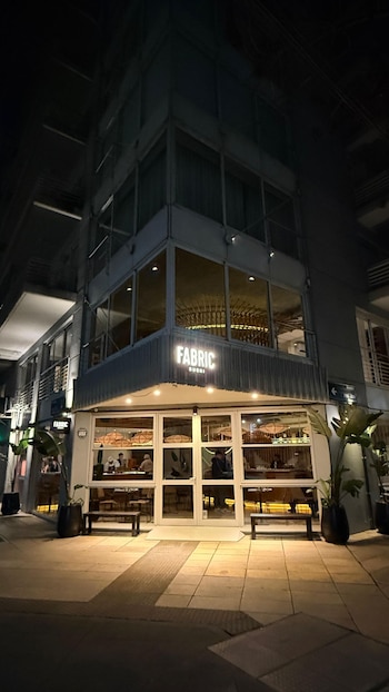 Esquina exterior de un edificio de noche con el letrero iluminado de Fabric Sushi, puertas de vidrio, mesas adentro y bancos en la acera