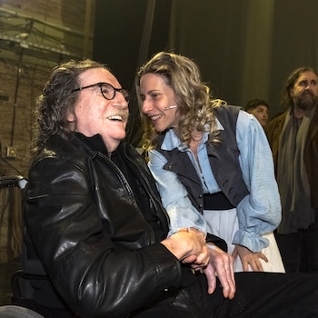 Charly García, un hombre con bigote, gafas y chaqueta de cuero negra en silla de ruedas, sonríe mientras Elena Roger, una mujer rubia con micrófono, le toma la mano