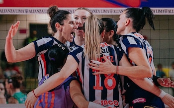 Jugadoras de voleibol con camisetas blanquiazules y de líbero morada se abrazan sonrientes junto a la red