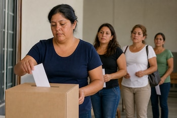 Una mujer introduce un sobre en una urna de cartón mientras tres mujeres esperan detrás con papeles en sus manos.