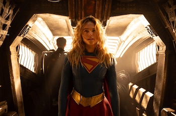 Una mujer vestida como Supergirl, con traje azul, falda roja, capa roja y cinturón dorado, se encuentra en un pasillo oscuro con otro hombre detrás