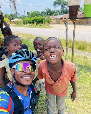 Un hombre con casco de ciclismo y gafas de sol se toma una foto junto a un grupo de niños cerca de una carretera rural