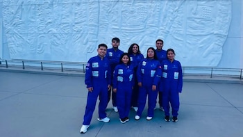 Siete personas, cinco mujeres y dos hombres, con monos azules, insignias de NASA y bandera de Perú, posan frente a un muro blanco con relieves y una barandilla
