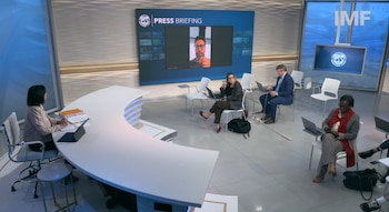 Una mujer en un escritorio curvo habla frente a reporteros y una pantalla grande con una videollamada en un estudio de televisión
