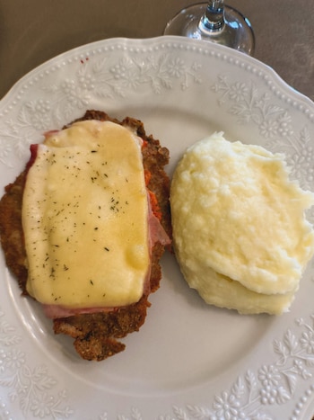 Un plato blanco con borde en relieve floral contiene una milanesa napolitana con jamón, queso gratinado y hierbas, junto a una porción de puré de papas. Se observa una copa
