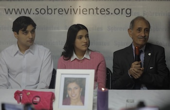 Un hombre, una mujer y otro hombre sentados frente a un banner con texto. Sobre la mesa hay un retrato de mujer, una vela y una bolsa rosa