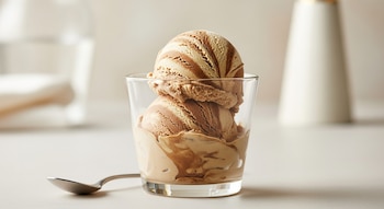 Helado de café y leche de almendras en un vasito transparente sobre una superficie clara, con una cuchara de metal a la izquierda.