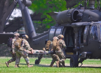 Cinco soldados en uniforme de camuflaje transportan a una persona en camilla hacia un helicóptero militar gris con la puerta abierta, en un campo de hierba