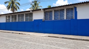Una fachada de edificio con sección inferior azul y superior blanca, ventanas con rejas blancas, techo ondulado, cableado, palmeras y cielo azul.