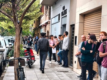 Larga fila de personas de distintas edades en una acera. Edificios al fondo, vehículos estacionados en la calle y un árbol grande en primer plano