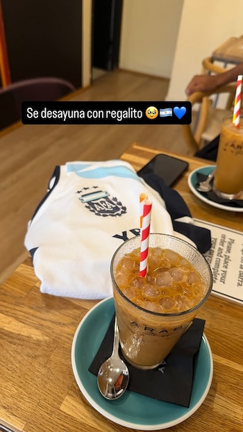 Una camiseta de fútbol de Argentina doblada sobre una mesa de madera, junto a dos vasos con café helado, pajitas de colores y una cuchara