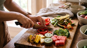 Manos que sostienen un cuchillo para cortar un pimiento rojo sobre una tabla junto a trozos de piña, kiwi, sandía y brócoli.