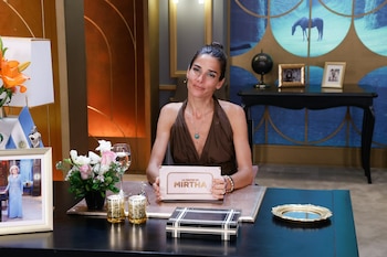 Juana Viale sentada ante un escritorio con un cartel del programa La noche de Mirtha, arreglos florales y un retrato enmarcado