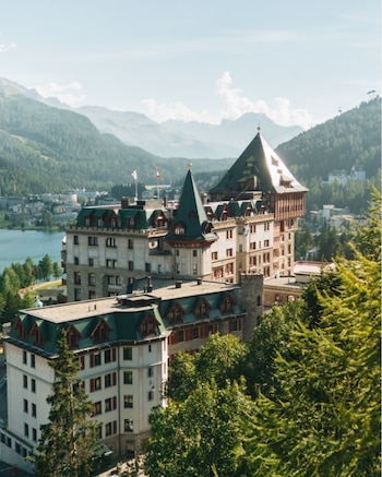 Edificio de varias plantas con tejados verdes y torres, rodeado de árboles. Al fondo, un lago y montañas cubiertas de vegetación. Cielo azul claro