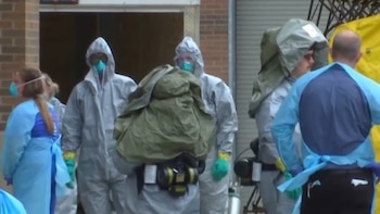 Múltiples personas con trajes protectores blancos, mascarillas azules y guantes, algunas con mochilas y equipo, frente a un edificio de ladrillo y una lona amarilla