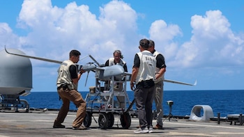 Cuatro hombres en chalecos trabajan alrededor de un dron gris sobre un carrito en la cubierta de un barco. El mar azul y el cielo nublado son visibles al fondo