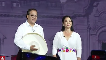 Keiko Fujimori y Roberto Sánchez de pie en un escenario morado, Fujimori con vestido blanco y fajín floral, Sánchez con camisa blanca y sombrero en mano