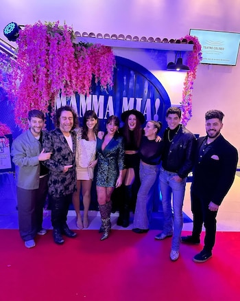 Ocho personas sonrientes posan sobre una alfombra roja frente a una pared azul decorada con flores rosas y la inscripción Mamma Mia; un televisor muestra Teatro Coliseo